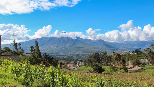 Small colonial homes dot green fields under tall Andes mountain range on a sunny day in Ecuador.