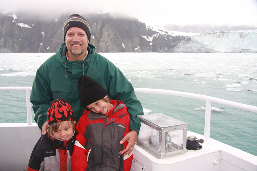 A father and his two sons seen on the bow of a boat in Alaska with a glacier behind them on a cloudy day