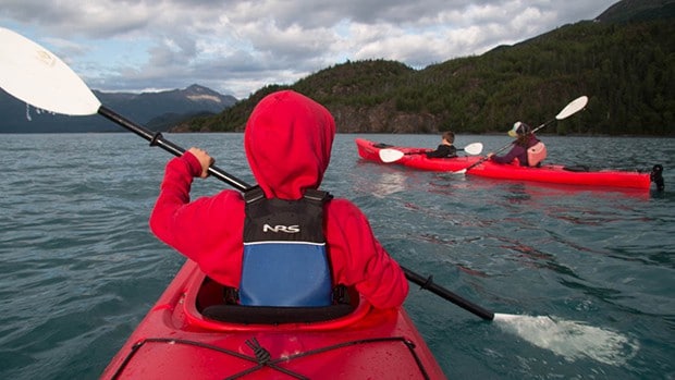 A kid in a red hoodie seen from behind kayaking in Alaska with another red kayak in front of him