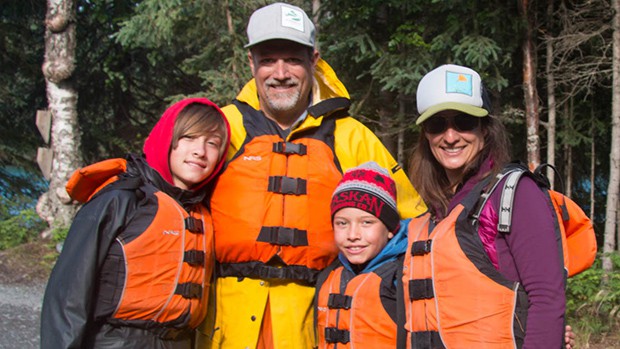 A family of four in life jackets and red, orange and purples jackets smiles looking at the camera