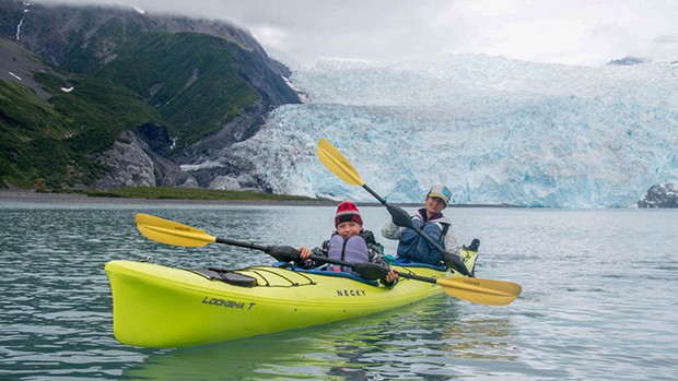 A mother and son in a yellow kayak paddle in front of a bright blue glacier in Alaska