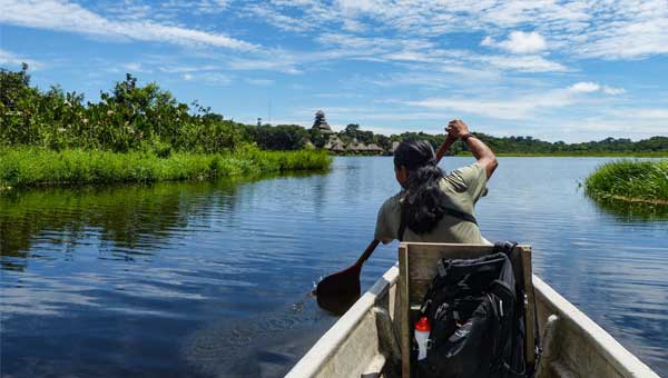 Man paddles dugout canoe across lake to remote Amazon rainforest tour eco lodge on a sunny day.
