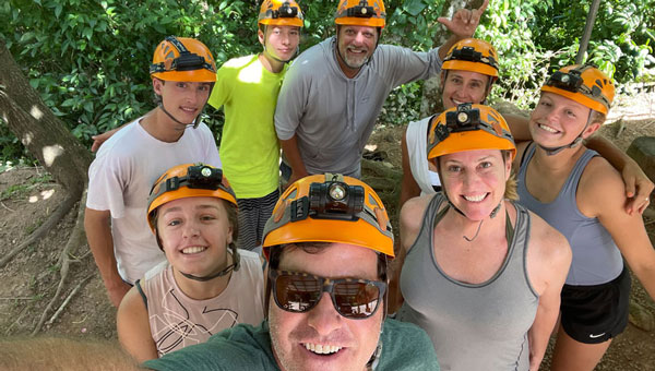 Kids and parents smile for a photo while wearing orange helmets with headlamps among forest on a Belize adventure.