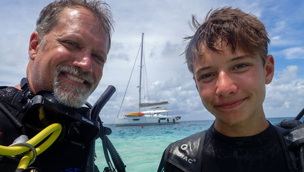 Man & boy in neoprene wetsuits & scuba gear stand in turquoise sea under sunny skies with white boat behind during a Belize adventure.