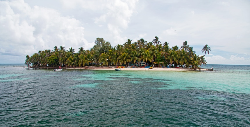 In Belize tiny sand island covered in bright green palm trees has colorful swing sets and chairs set along the shoreline,
