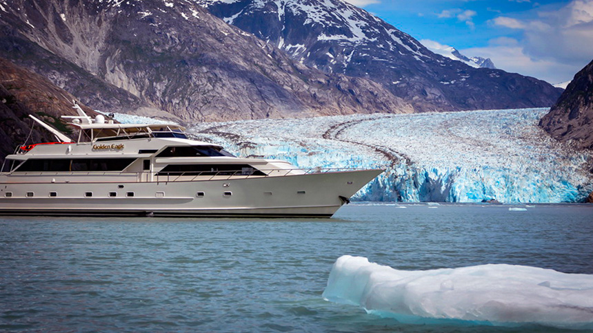 A small luxury Alaska cruise ship is seen at anchor in front of a big tidewater glacier with an iceberg in the water in front of the yacht