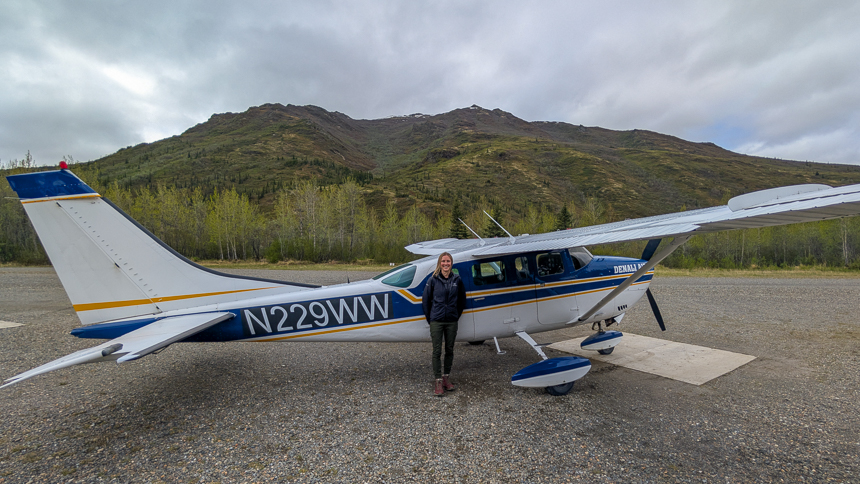 Women standing in front of small aircraft with blue and yellow strips in front of green mountains on a cloudy day
