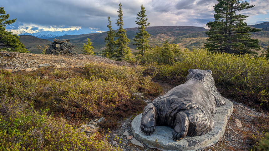 Statue of a bear lying on its belly over looking the mountains with pine trees and cloudy skis