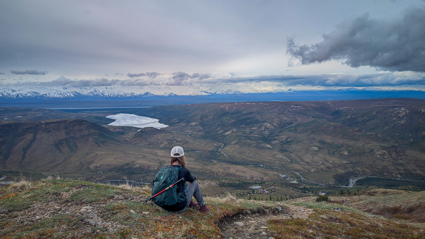 Hiker sitting with her backpack on looking out over lake wonder and camp denali on a cloudy day