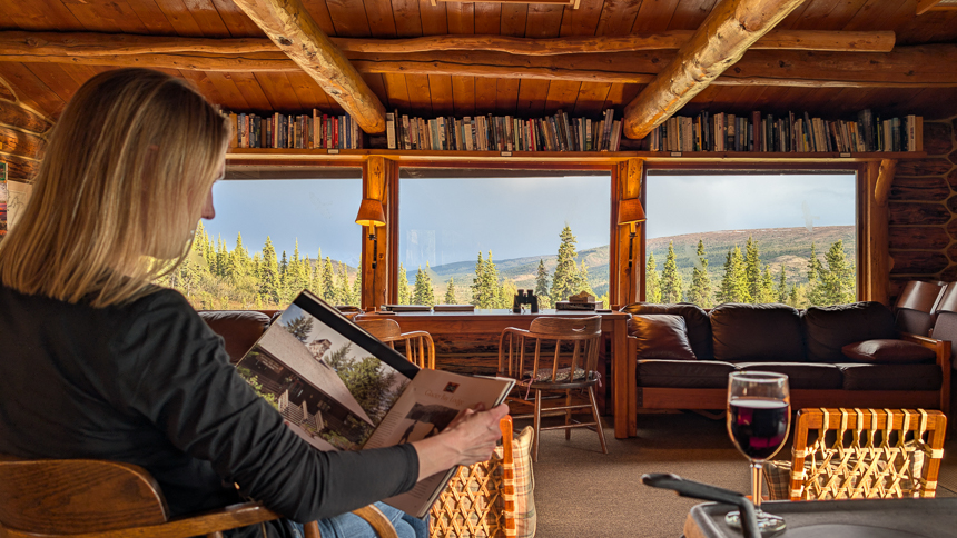 Women sitting in Camp Denali lodge reading a book and drinking a glass of wine looking out through the windows to the pine trees and mountains