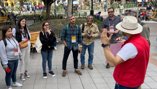 Man in red vest & white woven Panama hat talks to an Ecuador tour group in a city park.