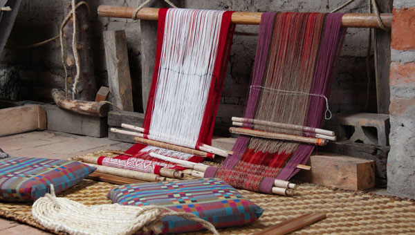 2 colorful pillows sit on stone tile floor in front of long wooden dowel with 2 hand weaving projects partly done in Ecuador.