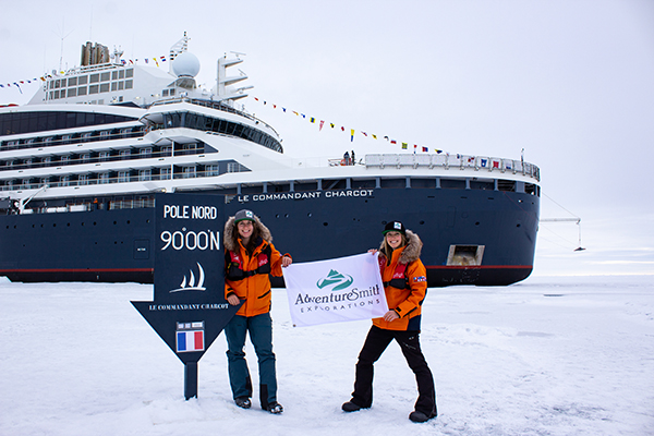 Two female travelers in yellow jackets stand in front of a small ship in sea ice with a North Pole sign beside them