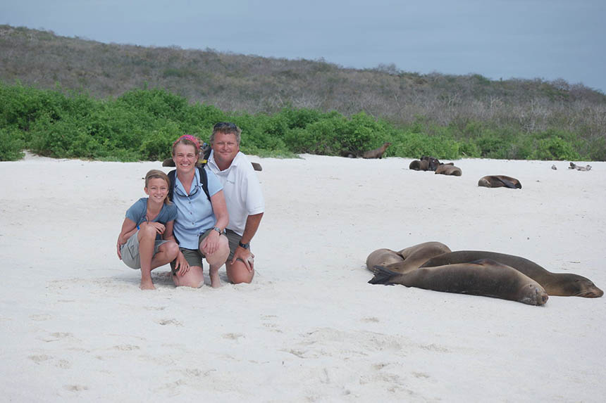 A family of three squats on a white sand beach next to Galapagos sea lions resting