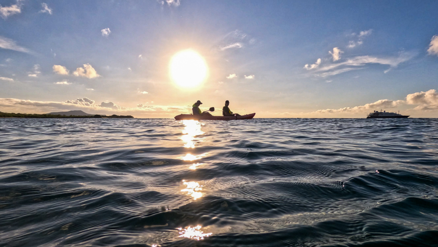 Kayakers paddling out as the sun sets off of a small ship in the Galapagos as the sun reflects off the water  