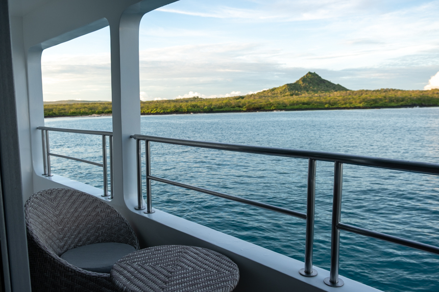 view from small ship infinity balcony with wicker grey seat overlooking a green landscape with a mountain 