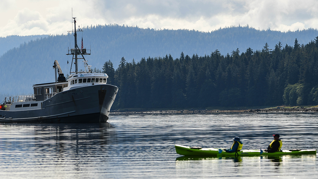 Starboard exterior of the Kruzof Explorer Alaska small ship, anchored on a cloudy day with a single kayak in the water in front of it