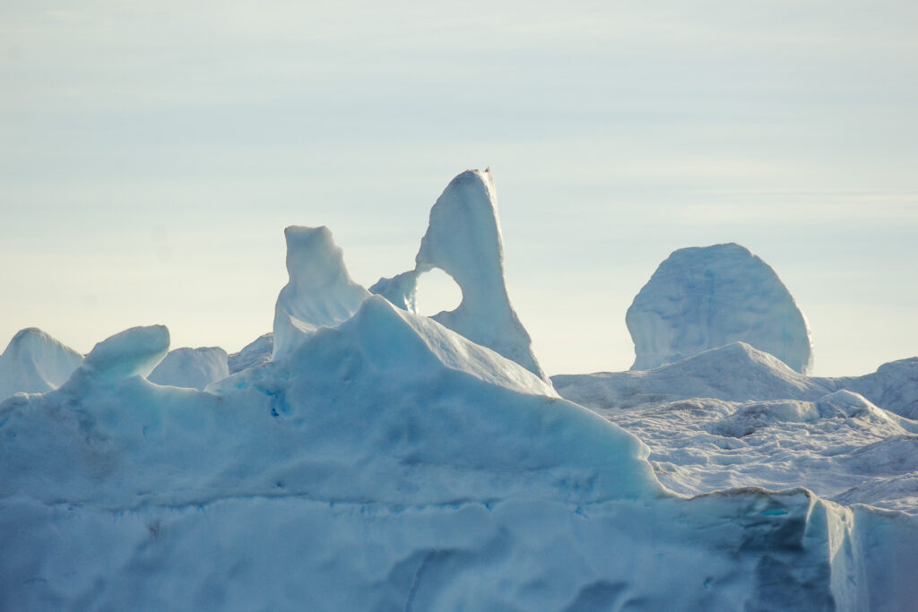 Large iceberg with unique ice melt cut-out contrasts against hazy mid-afternoon sky in Northeast Greenland.