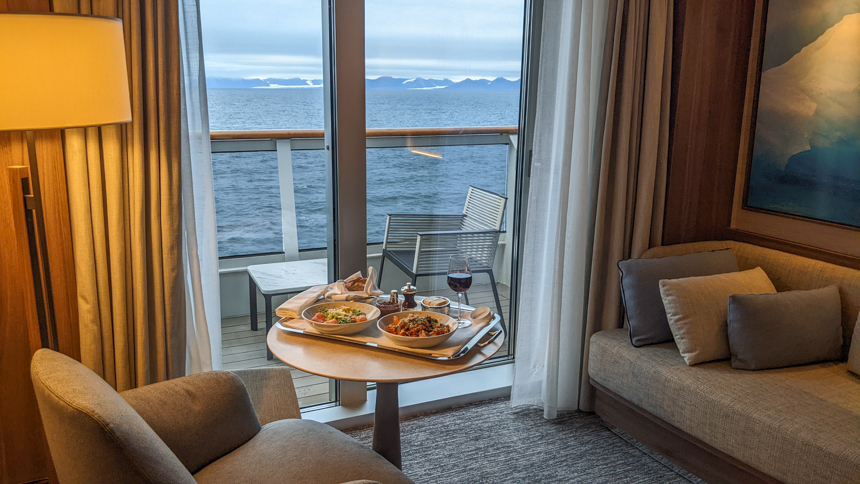 Room service food set on table in seating area of Deluxe Suite aboard Le Commandant Charcot with views from glass balcony doors