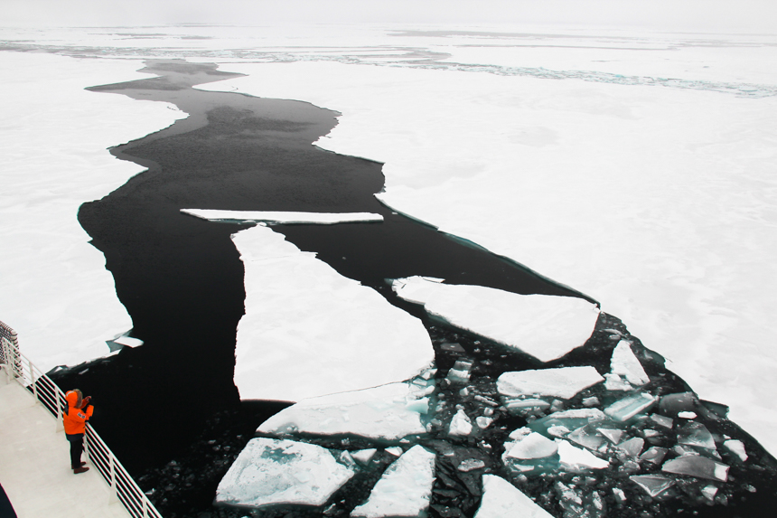 broken slabs of white ice float past the ship as it pushes through ice sheets. A traveler in an orange parka watches from the deck.