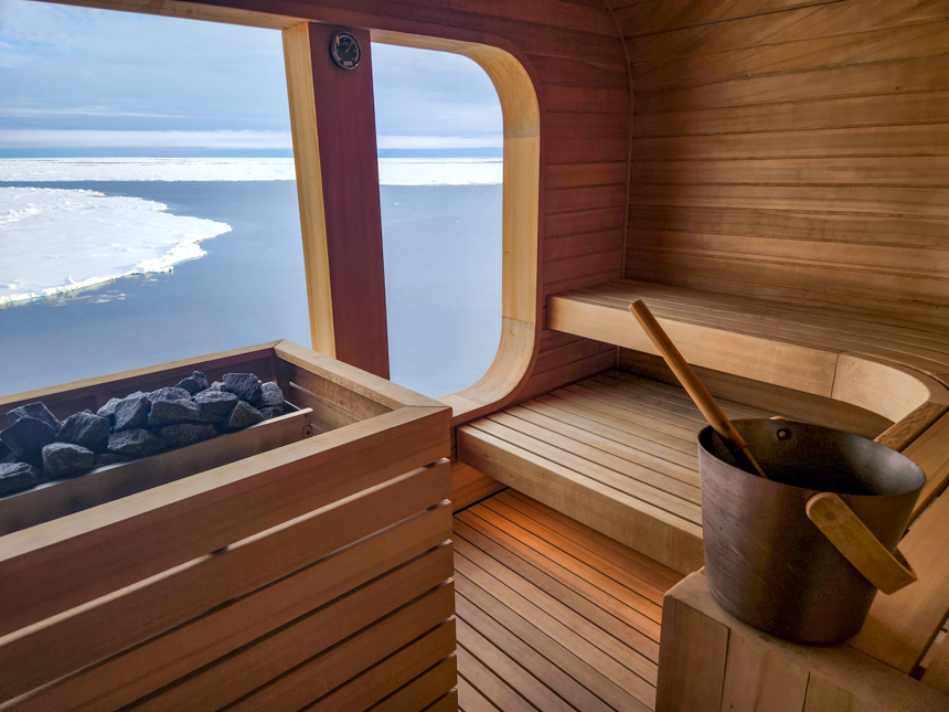 Indoor Wood Sauna with glass windows to the outside landscape, wood seating and black heater Stones with bucket of water.