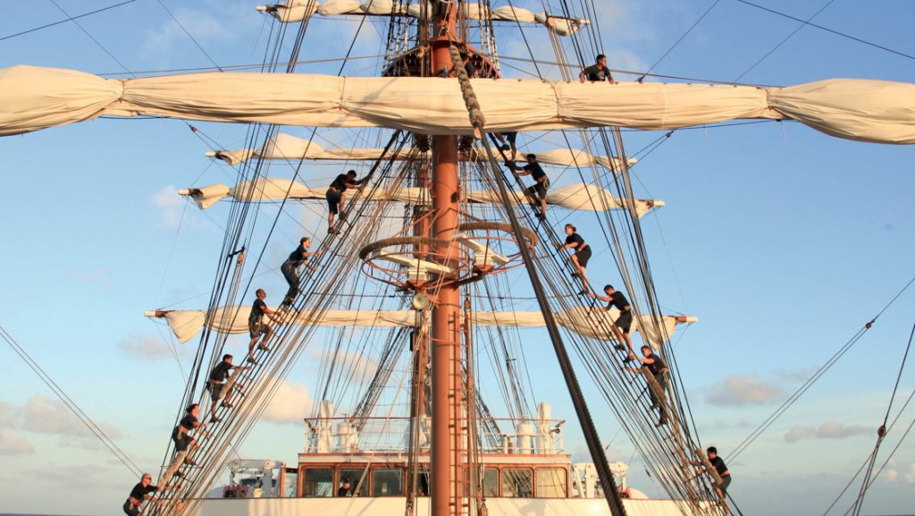 Crew members of Sea Cloud II: Lindblad ship climb the rigging to unfurl the sails with a calm blue sky behind.