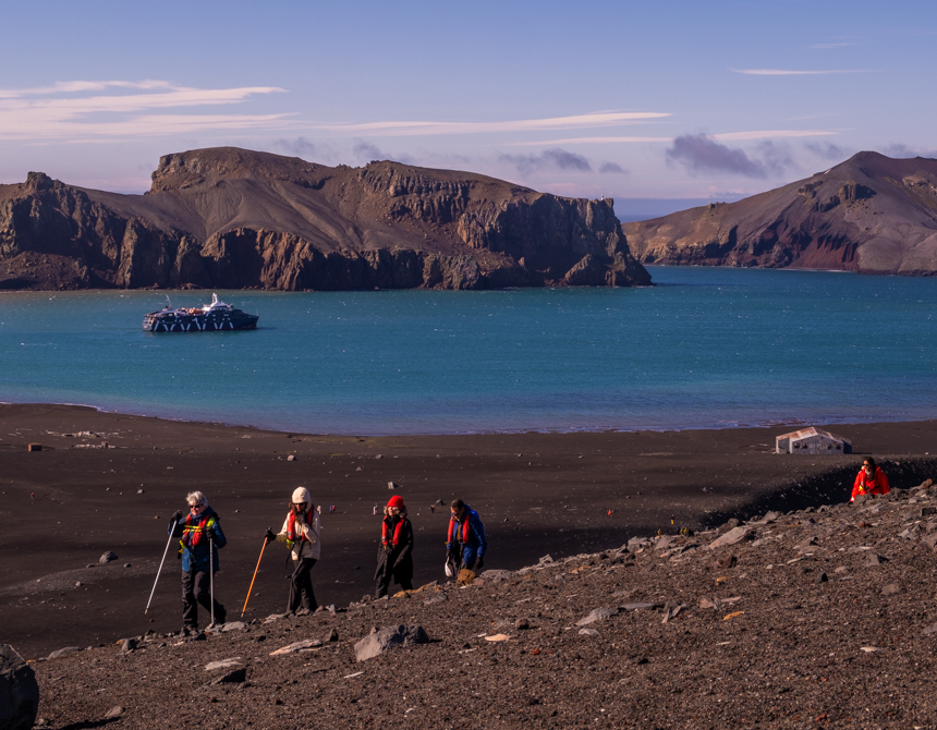 Guests with walking poles on Deception island in Antarctica on a sunny day with Magellan Explorer off in the distance