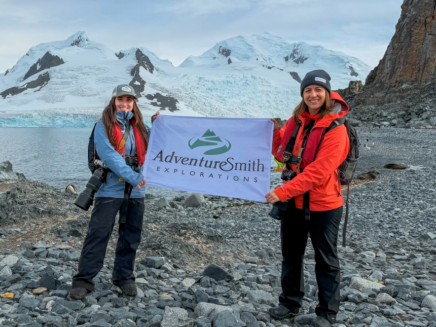 AdventureSmith Grace and Kelly are pictured with an AdventureSmith flag on land in Antarctica with seals and penguins off in the distance