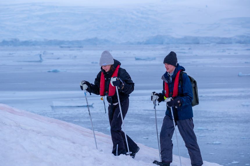 Guests with walking poles hiking up a snowy hill in Antarctica on a cloudy day with the ocean and icebergs off in the distance