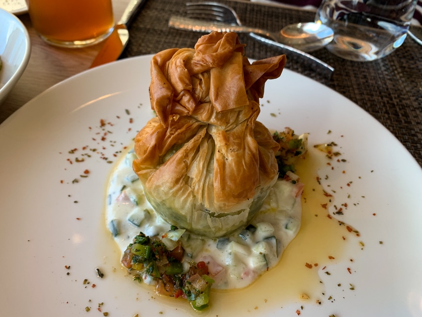 White dinner plate with creamy vegetables surrounded in oil and topped with a filled crunchy wrapped dumpling.