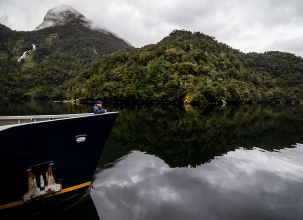 A man stands alone on the bow of a small New Zealand cruise ship seen with green trees and mirrored water