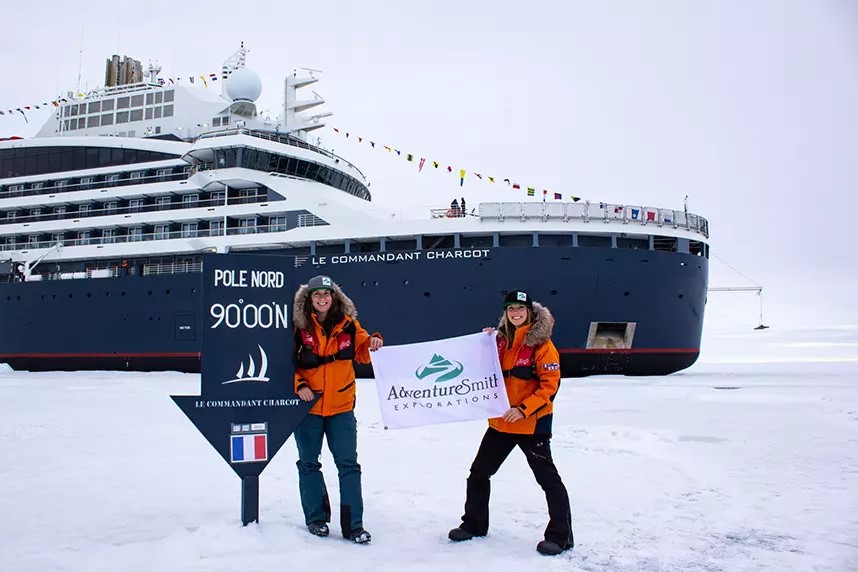 Two travelers in orange jackets stand at the North Pole 90 degrees blue sign in front of an expedition ship and holding an AdventureSmith Explorations flag between them
