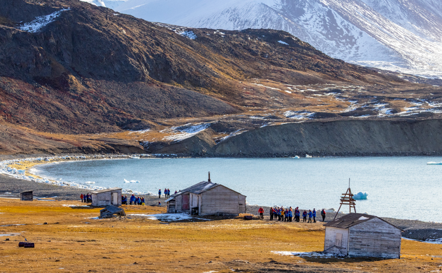 group of travelers walks through historic shacks on the Arctic coast on a bright sunny day