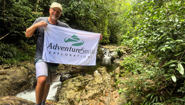 Man in shorts, t-shirt & hat holds a white flag with the AdventureSmith Explorations logo while standing beside a river with small waterfalls during a Panama tour.
