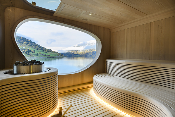Wooden sauna with 2 benches, hot stones & oval window looking onto mountains & water from a Ponant small ship.