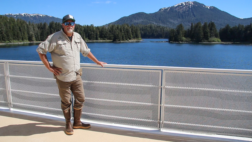 Man wearing tan safari shirt, tan pants and brown rubber boots stands with hand on railing of Safari Endeavor ship in Alaska.