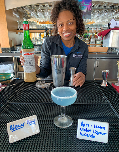 A bartender holds liquor bottle and bar tools behind a drink she just made, a icy blue cocktail called Glacier Bay