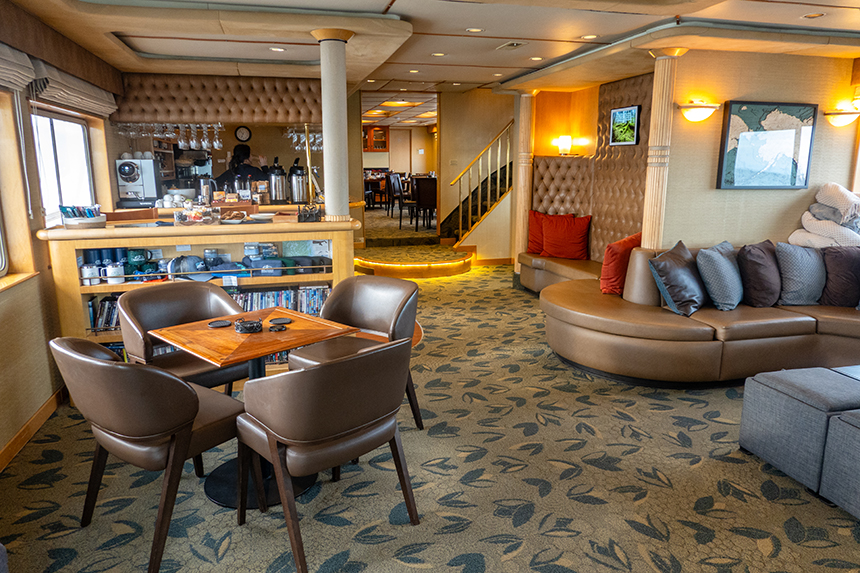 Cozy saloon aboard Safari Quest. Brown leather cushioned seating, pillows throughout, DVD shelf, bar, coffee table and ottomans.
