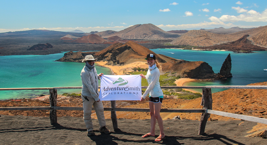 Male and female hold AdventureSmith Explorations logo flag at the top of a vista point overlooking beautiful Galapagos island volcanic landscape.