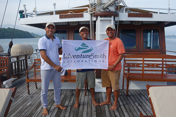 Three Indonesian men with barefeet hold a white AdventureSmith flag on the bow of a small ship cruise