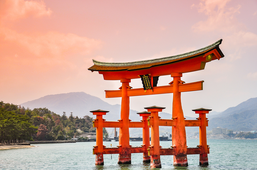 Red torii gate with Japanese characters stands over high tide in calm sea under a rosy sky at dusk on a Japan expedition cruise.