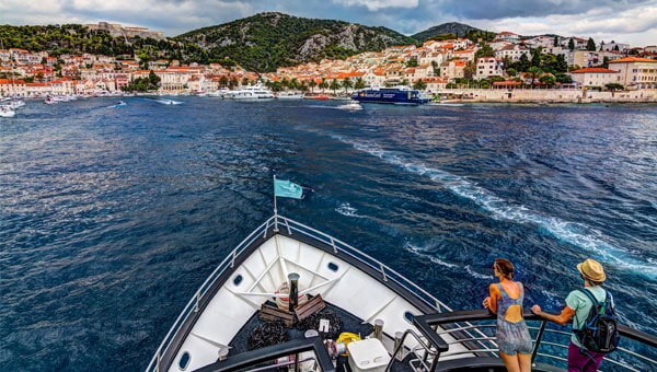 Two travelers on the bow of a white small ship cruise head towards a coastline in Croatia with red roof white homes and a green hillside