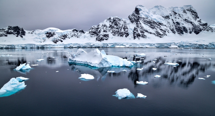 snowcapped icebergs floating in calm, grey ocean water in antarctica with snowcapped mountains in the background