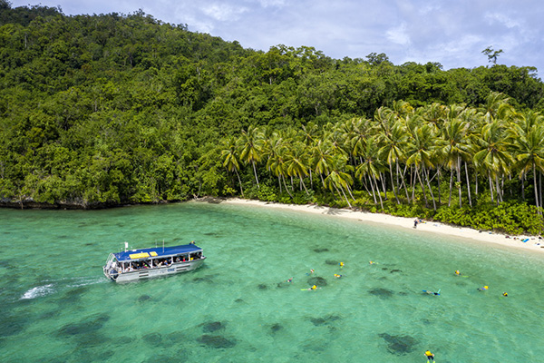 A small ship sails towards a tropical shoreline in Papua New Guinea with snorkelers in the clear green waters and lush palm trees on the shore