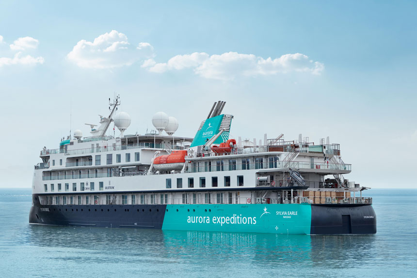 Sylvia Earle ship sitting in calm water, with blue & teal exterior with star logo, 5 white upper decks & 2 orange life boats.