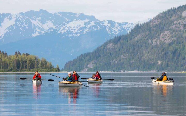 adventure travelers in yellow kayaks sitting in still water in front of an alaska mountain range