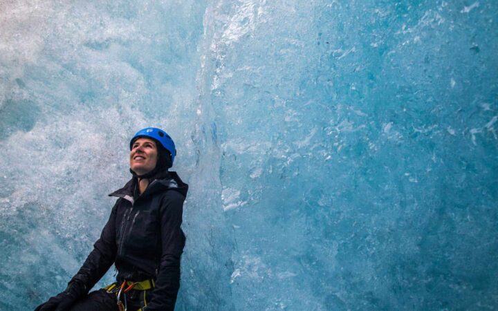 woman standing in front of an alaskan glacier wearing a helmet and smiling