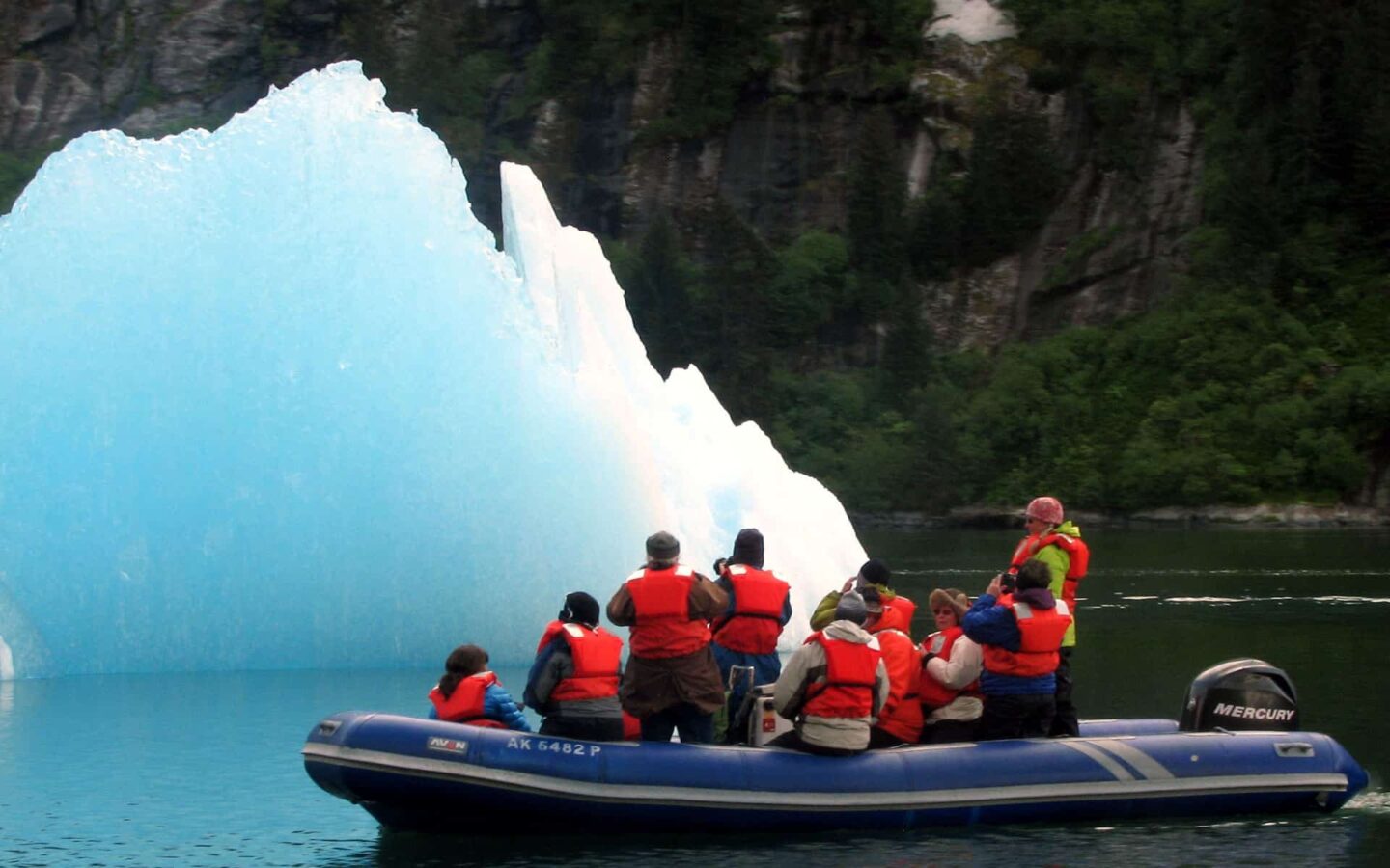 adventure travelers wearing red jackets sit in a skiff inflatable boat looking at a bright blue alaskan iceberg