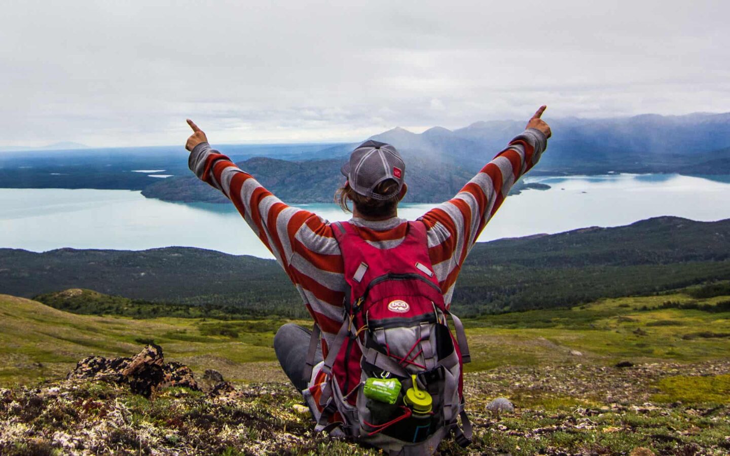 looking beyond a traveler with his arms in the air at an alaska inlet with mountains in the distance on an overcast day in alaska