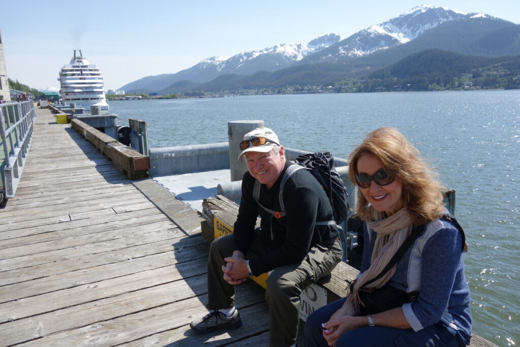 Two travelers sit on the waterfront dock in Juneau with a big ship behind them